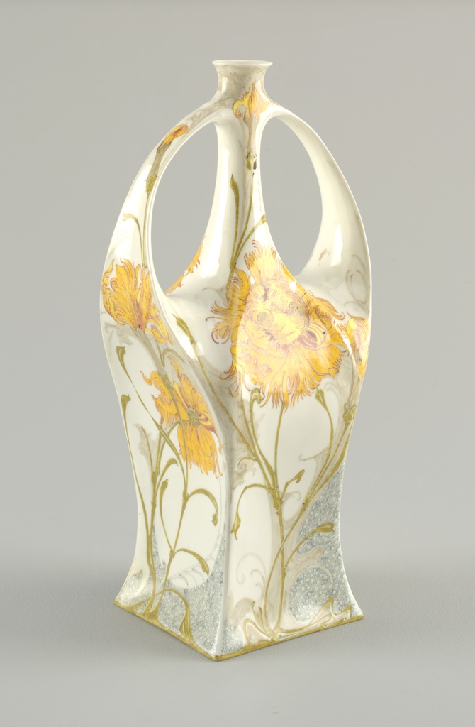 A tall white bottle. The central section contains a vase-like shape. On either sides of the top part are two handles, curving from 2/3 of the way toward the top of the bottle and up to meet the hole in the center. The design is floral. Narrow green stems blossom from the base of the of the bottle and curve upward. Yellow pedals appear at various places.