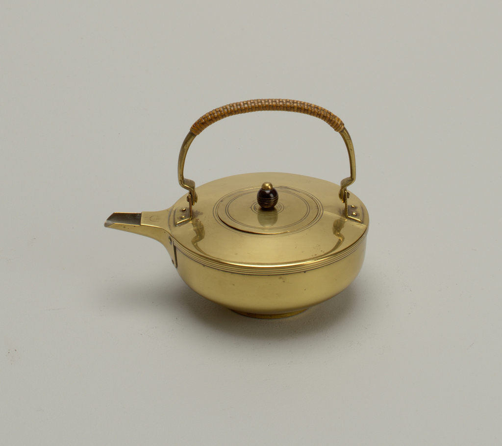 A brass tea kettle, with a squat, ovoid-shaped body and a short spout pointing to our left. The kettle has a curved, tapered handle attached to each edge with brass rivets, the central section of which is covered with an applied copper colored strap. In the center of the body is a small circular lid, engraved with two concentric lines, which has a small, dark brown, glazed wooden, spherical finial with a brass top, that is surrounded by two more engraved concentric lines.