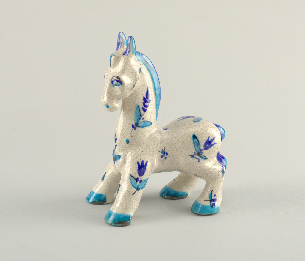 A small glazed earthenware horse figurine in left-facing profile. The mane, tail, ears and hooves are glazed in royal blue which fades into a bright turquoise, while the body is an off-white crackle glaze and is littlered with small deep blue flowers with turquoise leaves and black stems. The visible eye is bright turquoise with a royal blue eyelid, black pupil and a black arched eyebrow, while the nose is accented with a turquoise nostril.