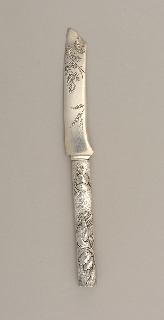 Sterling knife. The blade is shaped like Japanese sword with fronds engraved on it. There is a clipped end where the point would be and below that the blade curves slightly downward. The handle has a mottled surface with two leaves on branches on top and bottom and a bird in the center.