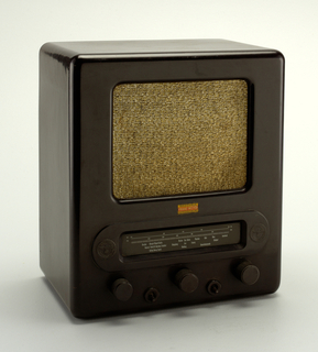 Here we have a brown, rectangular radio with curved edges and corners. On the front, there is a large square speaker covered with brown and black woven fabric. Below the speaker is an elongated station indicator window, with embossed circular eagle logo emblems on both ends. Below the station window are three circular dials and two small switches in between them.