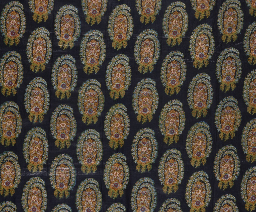 Dark black blue textile with offset paisley pattern in columns. The textile has a mild sheen. The pattern consists of two floral sprays wrapping around a central floral arrangement forming an oval. Small flowers in red and blue surrounded by green leaves line the exterior on a thin yellow branch, small buds line in the interior. The left spray wraps up and over the right. At center a symmetrical floral arrangement with various flowers in blue, green, red, and yellow. The bottom of the arrangement centers on a blue and red flower. A yellow stem extends from this flower with two short downward curved branches with red flowers on either side. The exterior floral sprays connect at this stem.