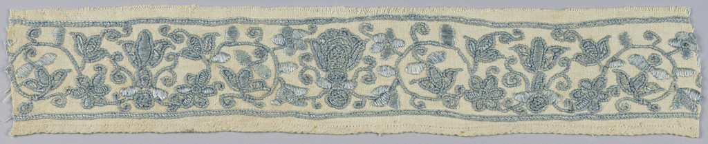 A horizontal band of cream-colored fabric embroidered with scrolling floral motifs in smokey, faded blue extending horizontally across. The band is narrow, and the embroidery occupies most of the surface. A line of blue frames the top and bottom of the floral pattern, which is stylized and repeating. The left and right edges of the piece exhibit slight fraying.