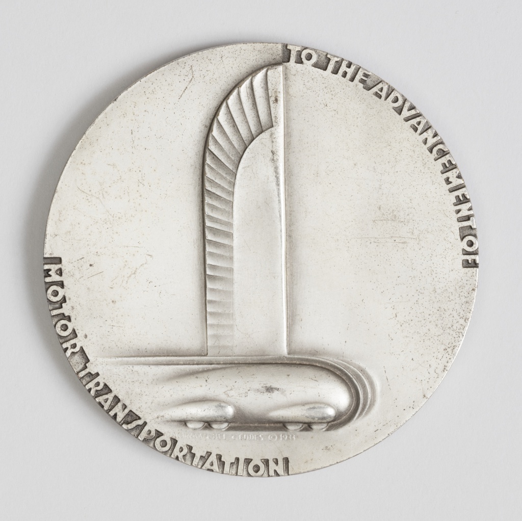 Round medal made out of silver-plated brass. Sections of the outer edges of the medal are embossed with text. “TO THE ADVANCEMENT OF” on the upper-right section and “MOTOR TRANSPORTATION” on its lower-left. At the center is an embossed rendering of a stylized car with vertical wings. At the bottom of this symbol is the text “NORMAN BEL GEDDES © 1933”