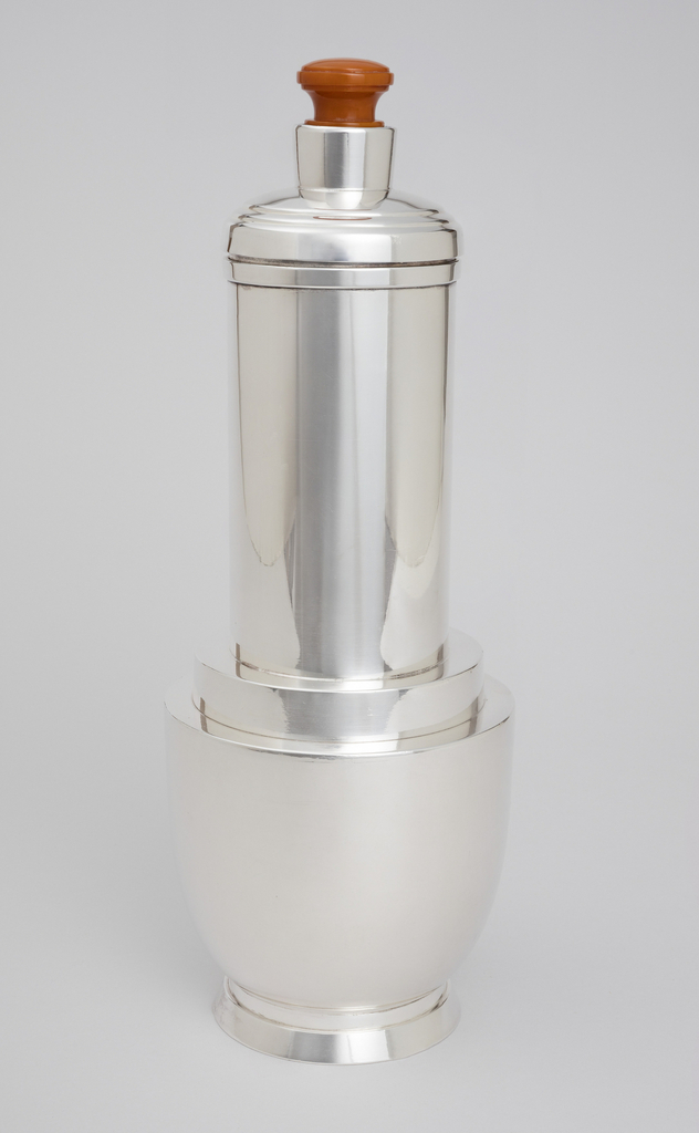 A mostly silver-plate cocktail shaker with a U-form dominating the bottom segment, also known as a spun hemisphere, with an angled base prone in an outward direction at the bottom. In an upward direction are two steps inward to a tall cylindrical body resembling a form of a skyscraper with two more steps out and three shallow steps inward leading to a lid with a neck and a red-orange Catalin (polymer) stopper that remains inserted during pouring. The photograph captures the cocktail shaker in a front view against a medium gray background.