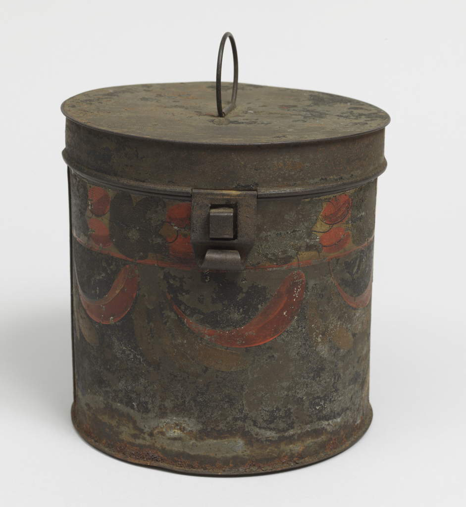 A dark brownish-black tôle (a metal alloy consisting of iron and tin) cylindrical box. The box has a round lid with a thin handle on the top in the center. On the front side near the top is a large hinge. The box has been painted with bands of olive green and rust-colored foliate decoration, including flowers at the top and swags in the center. The tôle is worn and shows dark-colored rusted sections across its body.