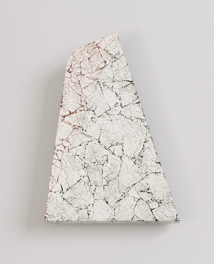 This four-sided brooch is shaped like a tall triangle with the top point trimmed off. The top left 'corner' is curved. The surface is smooth, inlaid with crackled pieces of white eggshell in varying sizes, creating a stone-like appearance. The spaces between the eggshell pieces and the fine cracks in the shells are colored dark red and black. These dark colors are split into two triangles, a dark red triangle on the left and a black triangle on the right. The dividing line runs between the top right and bottom left corners.