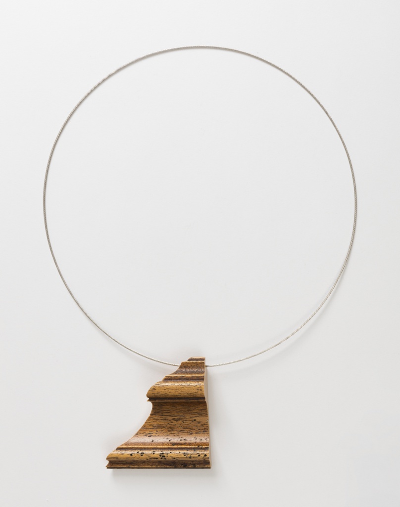 A circular necklace made from a thin steel cable with no visible clasp, with a pendant made from a triangular section of medium-toned wood picture frame molding, with undulating ogee curves, lateral grain and some wormhole texture.