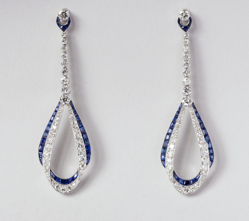 A pair of earrings in an abstract teardrop setting encrusted with faceted diamonds and rectangular sapphires set in platinum. The earrings begin as a diamond post where a small inverted sapphire teardrop form is suspended from the post. Hanging below is a line of round diamonds increasing in scale. Attached at the bottom is a large abstract teardrop form embedded with small round diamonds, overlapping a similar teardrop form embedded with faceted sapphire stones that appear to be calibrated to fit the space, in a stationary setting.