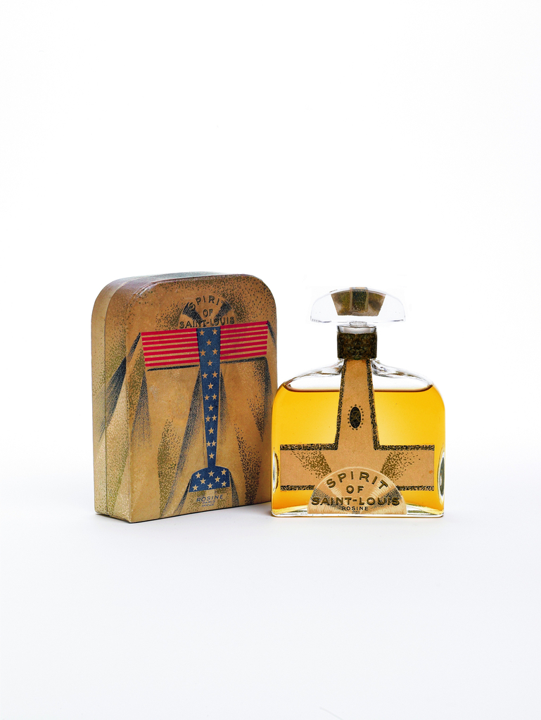 An airplane-themed perfume bottle and matching box on a white background. The bottle is clear glass, with a wide rectangular form, sloping shoulders, narrow neck, and wide stopper with a rounded top edge. The perfume within is a warm golden color. A light brown label in the stylized shape of an airplane – wide horizontal wings, narrow tapering vertical body – fills the front of the bottle, with the half-circle propeller at the bottom and the tail on the stopper. [SPIRIT OF SAINT-LOUIS ROSINE] is printed on the propeller, with whirring white accents suggesting its spin. Speckled black outlines accent the airplane. The matching box is a corresponding shape, rectangular with rounded top corners, also decorated with an airplane. This one faces up, with an American flag design of red-striped wings and a starry blue body and tail. [SPIRIT OF SAINT-LOUIS] is also printed in the spinning propeller, with [ROSINE] in italics below the tail. The aged brown box is marked with dynamic black-speckled wedge shapes angling away from the plane, suggesting wind streams in flight.