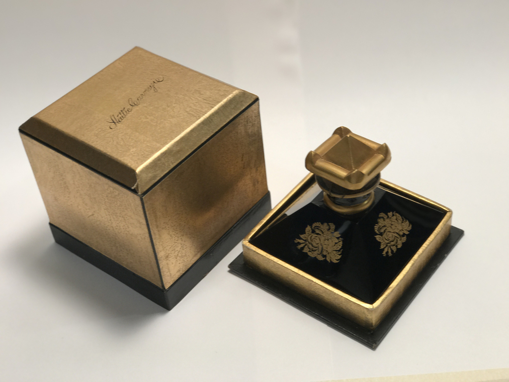 Here we have an elegant, pyramid-like perfume bottle with black glass enamel and accented gold gilding placed on a small, square black tray. The four sloping, black enamel sides emerge from the upright, low, gilded base. On the shiny black enamel is an intricate gold floral bouquet decoration with leaves, rose, and foliage forms. Around the top neck are two thin rolls of gold gilding that widen toward the cap. The cap is a unique pyramid shape with four pointed corners and is also gilded. Accompanying the perfume bottle is a square, gold box with a slanted top and cursive inscription [Hattie Carnegie].