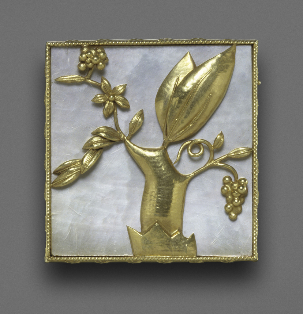 Small, square Wiener Werkstatte pin of mother-of-pearl, trimmed with ropelike twisted gold wire. Attached to the mother-of-pearl is a highly stylized gold sculpture of a tree, from which grapes, leaves, and flowers bloom. Two of the leaves are disproportionately large while the trunk is disproportionately short. The tree sits in what appears to be an inverted crown that is on a slant.