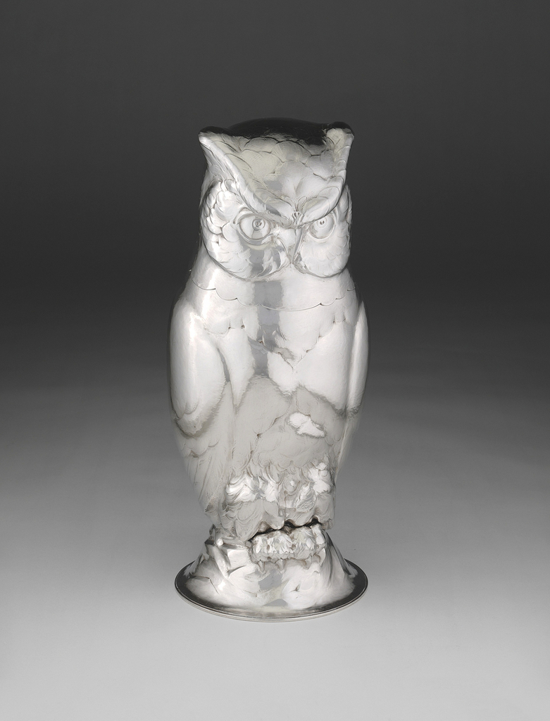 This owl looks remarkably like a silver sculpture but is in fact a cocktail shaker. Perched on a detachable base, the owl is sleek in design but fairly true-to-form in the animal's features and overall sculptural style. It is monochrome, has a smooth surface, and you can see layers of feathers on its head, at the bottom of its wings, and near its feet. There is a small indentation around its neck to suggest that is where you can remove the top and mix the drink in its body.