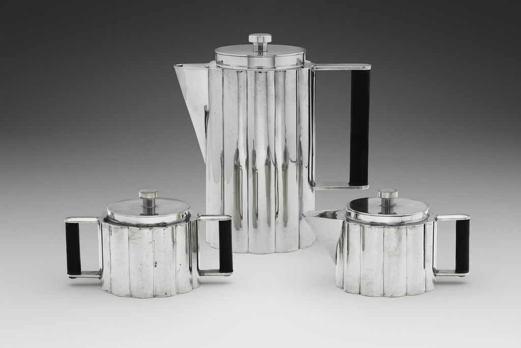 A three piece hand-wrought pewter coffee set. On the left is a double handled sugar bowl. The back middle showcases a tall standing pot with wedged lipped spout. On the right is on a squat cream server. All three are lidded with a shiny exterior and the interiors of the handles set in black. They are photographed against a cascading black to white background.