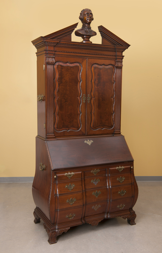 A large wooden hutch. At the bottom are small drawers, organized four by three (ie, four horizontal rows and three vertical columns). Each draw has a small brass handle. On top of the drawers is a small fold-out desk that slants outward from top to bottom. On top of the desk is a vertically rectangular cabinet with wood engravings on it. On top of the hutch is a bust of a male head.