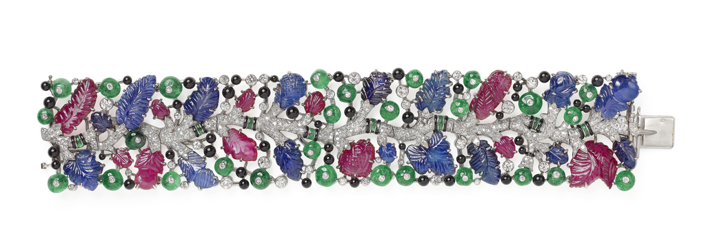 Lateral view of an oblong rectangular platinum bracelet with a flattened clasp, featuring a central winding vine of pave diamonds with occasional onyx and emerald cuffs and carved ruby and sapphire leaves and fruits emerging from either side, bordered by an outline of round diamond-centered emerald berries, small round onyx berries and round diamonds.