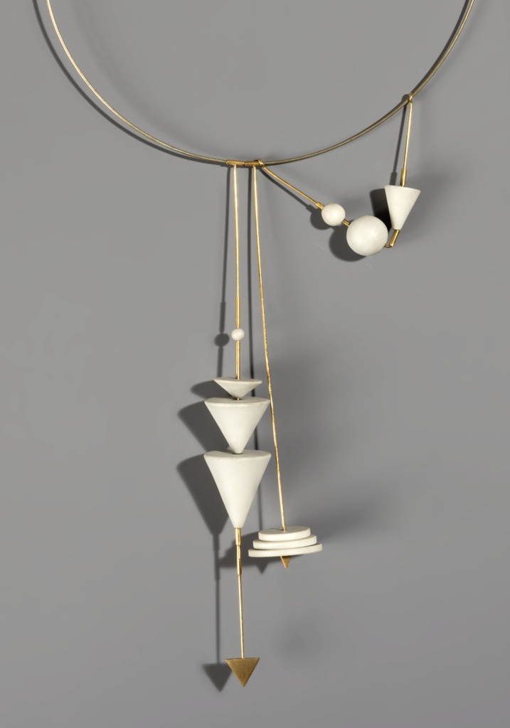 Partially displayed, a thin gold hoop necklace. Hanging from the hoop are separate forms; the first form is a equally thin gold beam which holds three upside-down white cones of various sizes, and and at the very end is triangle tip. The second form (to the viewers right side) is the same gold beam but a tad shorter, with three white discs stacked upon each other and a gold triangle at the end. To the right of that (according to viewers perspective) is the final accent form; two gold beams that connect together, creating a triangle. It contains two small but different sized white spheres and a singular cone.