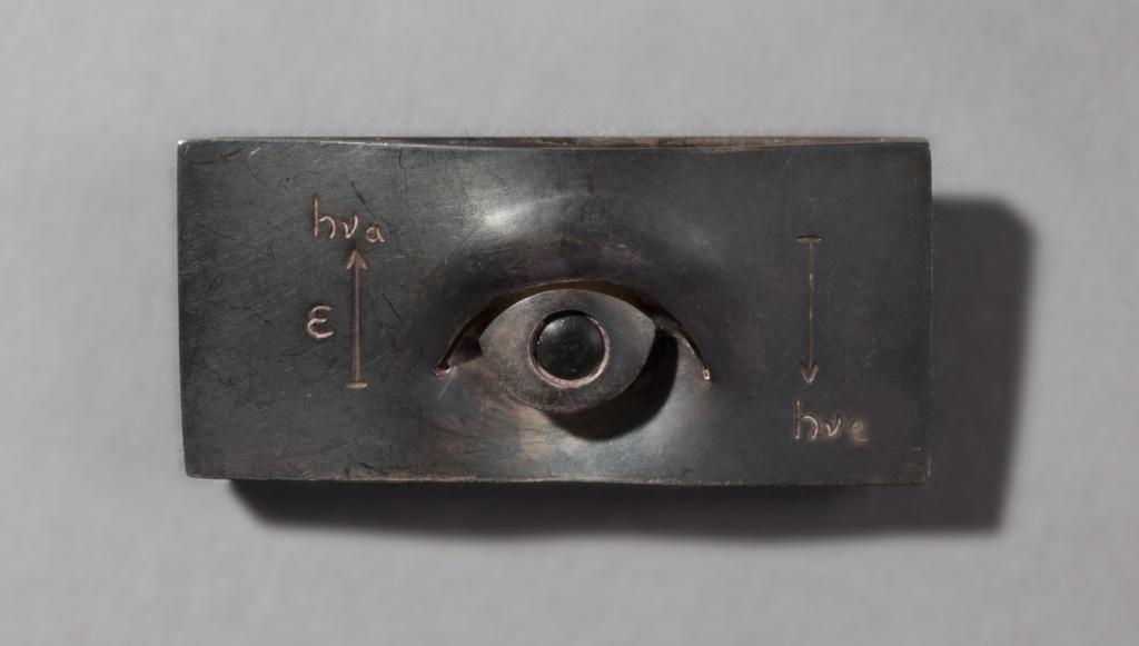 Brooch in a rectangular form made out of patinated and etched copper and stone. At the center is an embossed, relieved dimensional figure of an eye. Upper lids slightly raised and the bottom area has a curved groove. The sclera and iris are separated and raised creating the dimensional effect. The iris is etched and is filled in with black. Several text and symbols are also etched on the sides of the brooch. On the left, an arrow pointing upwards with the text “hva” on top of it. The letter “E” appears on its left. On the right side is an arrow pointing down with the text “hve” beneath it.