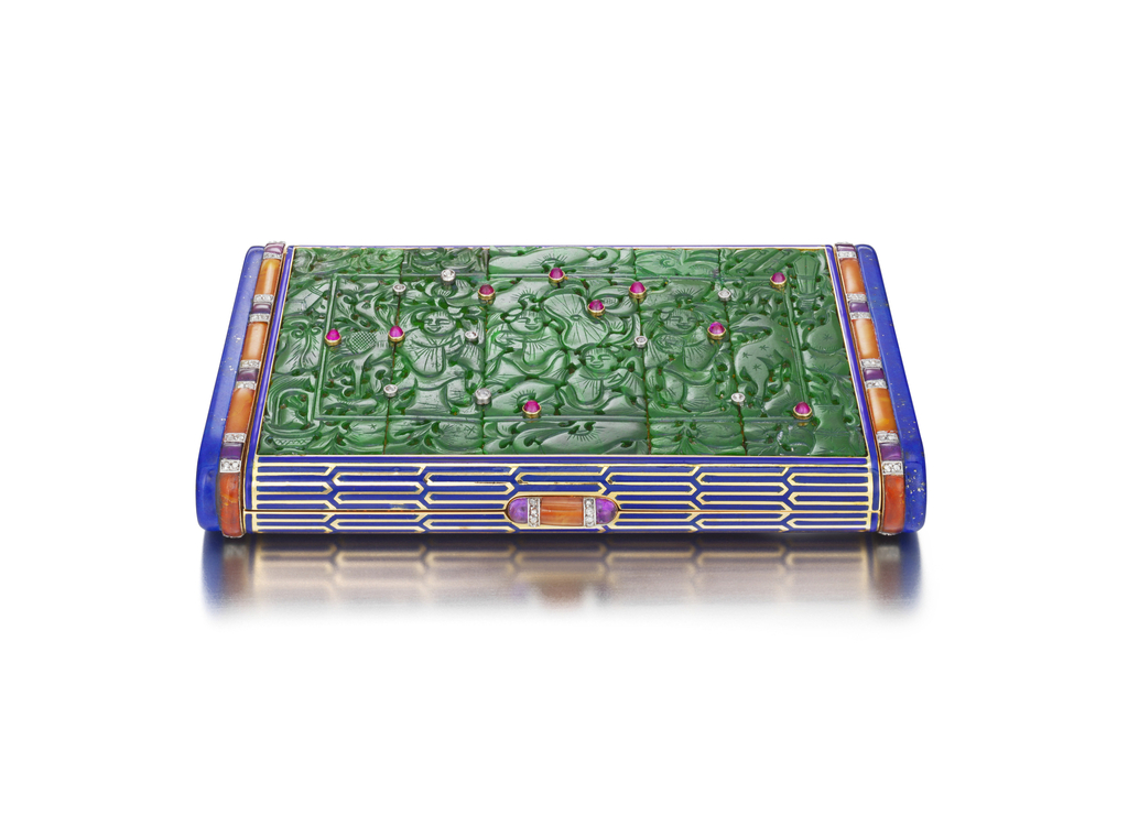 Rectangular vanity case composed of a colorful assortment of semi-precious and precious materials. The top is rich green carved jadeite plaques depicting four smiling figures surrounded by lush plants and animals: a bird at left and a deer at right. The scene is studded with diamonds and bright pink cabochon rubies in platinum and gold settings. A thin line of deep blue enamel encloses the scene, continuing to the curved front edge, which is decorated with an interlocking geometric design in deep blue enamel on gold. The sides feature striped bands of orange carnelian and purple amethyst interspersed with diamonds. The thumbpiece, centered on the front edge, is similarly decorated. The short sides are deep blue, gold-flecked lapis lazuli, the same color as the enamel.