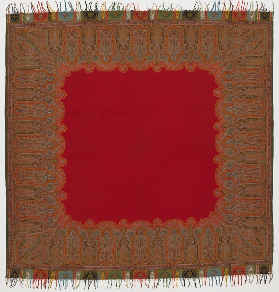 Shawl with frayed top and bottom. At the center is a red square with small wavy, yellow-lined border. Surrounding the square is a thick, brown/dark-green pattern. The pattern consists of two teardrop shapes facing each other, with a smaller, circlular shape at their meeting point. This pattern repeating about seven times per each side of the square. In between each pattern is a flower-like shape, with lots of small petals blooming in the opposite direction of the meeting point of the two teardrops.