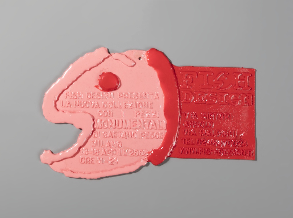 A horizontally placed form in low relief of a playful fish in profile composed of pink and red resin. The cartoonish fish's head is designed in pink resin with a red resin raised dot for the eye and far left, a raised elongated S line that emphasizes the opening of the fish's mouth. A tiny hole is included at the top of the head for a possible nail. The fish's body, right of center, is a red rectangular invitation embossed with the same color lettering identifying the name of the object/exhibit, Fish Design, and the gallery and address in Milano, where the exhibit was held. The stylized S in the word Fish, as in Fish Design, mirrors the S form of the object's profile or left edge. The designer's name, Pesce, means fishery in Italian, hence the probable choice of imagery through the art.