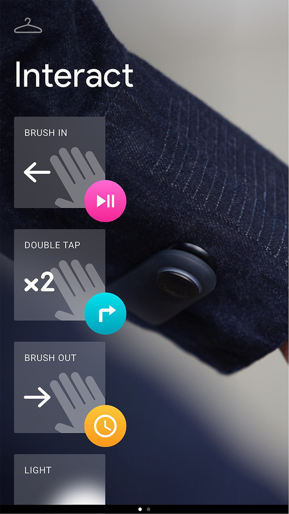 A screenshot from an app, showing a shirt sleeve and hand gesture controls within a vertical rectangle. The background image is of a dark denim shirt sleeve, with horizontal white stripes and a black plastic tag at the cuff. In the top left corner of the screenshot is a grey icon of a clothes hanger. At upper left is white text reading 'Interact'. Below are square boxes along the left side of the screen, showing greyscale icons of hands with white gesture text and indications. Each box has a colored icon circle at the lower right corner. First, 'BRUSH IN' with a left arrow; in the bright pink circle is a play/pause icon, a triangle with two vertical lines. Second, 'DOUBLE TAP' with 'x2'; in the bright teal circle is a right turn arrow. Third, 'BRUSH OUT' with a right arrow; in the golden yellow circle is a clock icon. Fourth, 'LIGHT' and a white glow. This box is cut off by the bottom of the screen. At the bottom edge of the screenshot is a white dot next to a grey dot, indicating another screen is available.