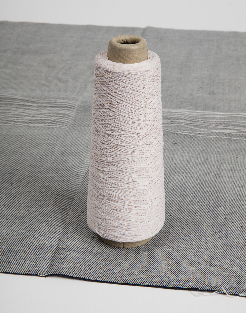 A tall, upright spool of white thread wound around a brown cardboard bobbin. The spool sits on a piece of heavy woven grey cloth, like the backside of denim.