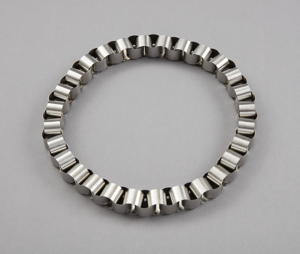 A laying flat circular necklace made of interlocked small cylindrical steel links.