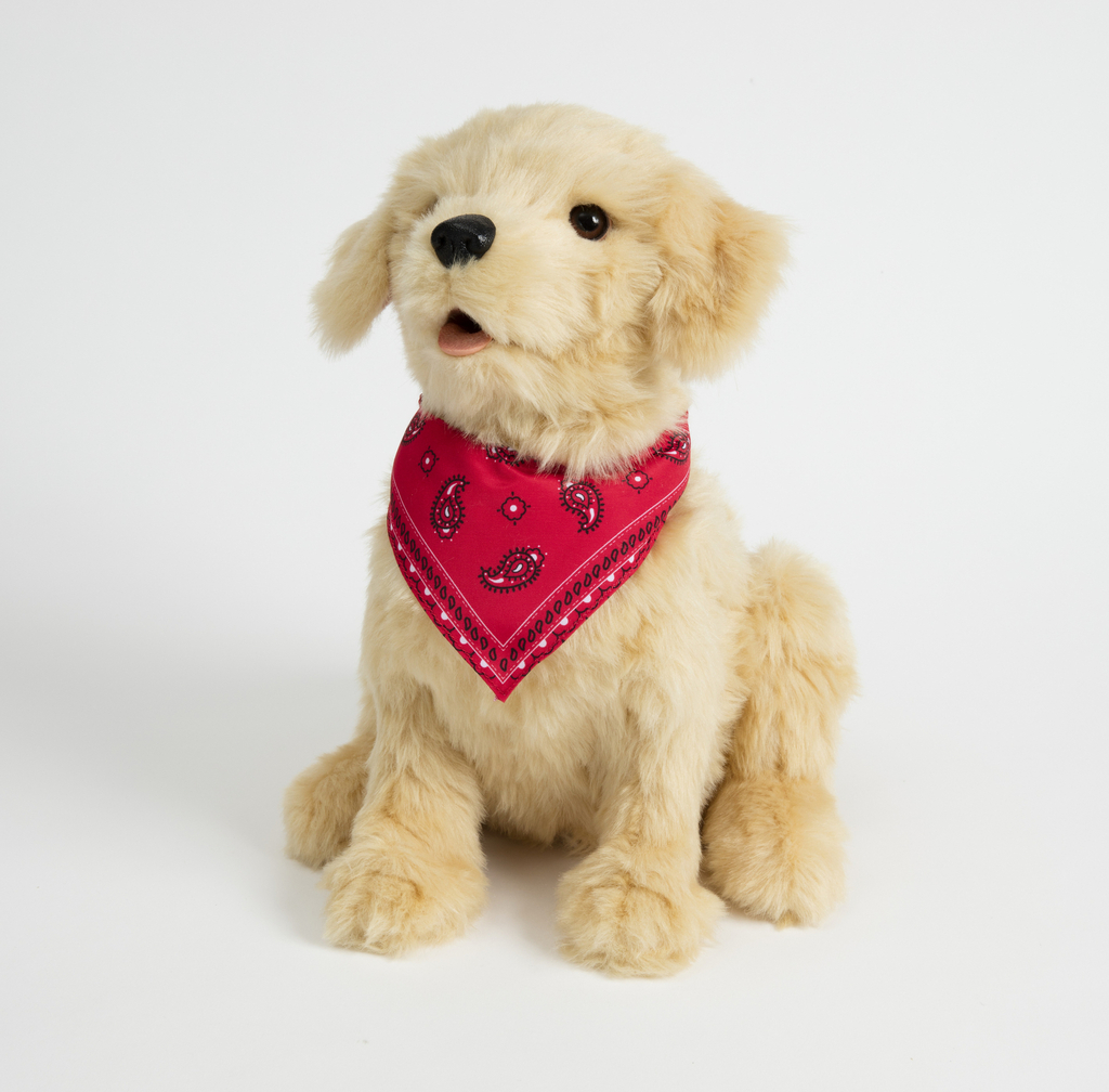 This small stuffed animal dog is sitting down with its head tilted upward and tongue partially sticking out. It has short, small ears, and black nose and dark eyes. It is reminiscent of a golden retriever puppy, with light, fluffy, golden-brown fur. It has a red handkerchief around its neck with black and white paisley decorative pattern.