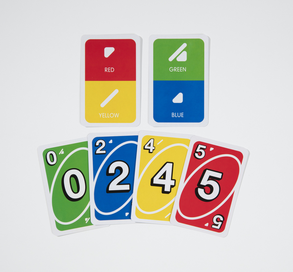 A set of six playing cards for the card game UNO which are split into two rows. The top row has two cards which are both split in half, the first red and yellow, and the second green and blue. These cards also have the symbols for color blindness printed on them and the word corresponding to the color. The red half has a right-facing, right-angled, white triangle, and the word [RED] in white capitals; the yellow half has a white, diagonal line, and the word [YELLOW] in white capitals; the green half has a white, diagonal line with a small, left-facing, right-angled white triangle below, and the word [GREEN] in white capitals; and the blue has a left-facing, white, right-angled triangle, and the word [BLUE]. The bottom row has four cards which are blue, with a number 0 on it; green with a number 2 on it; yellow with a number 4 on it; and red with the number 5 on it. Each number is encased in a eliptical shape with a white border and has a smaller version of the number in the top left corner; the red card also has a number 5 in the bottom right corner.