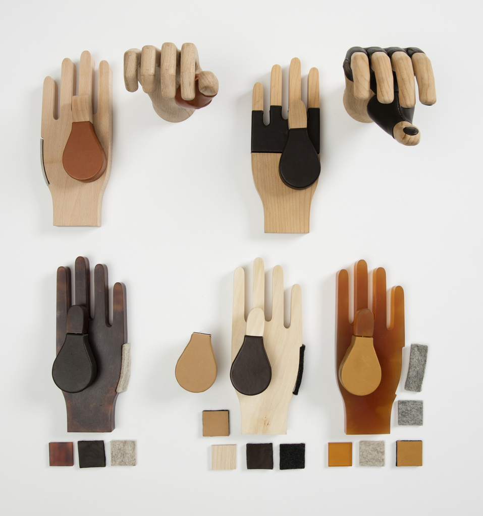 On a white background, seven hand prosthetics composed of varying materials is displayed in varying positions. On top are four of the hand displays, beginning with two light brown hands and a dark brown ball of the thumb. One is displayed palm down and the other, a top view with fingers curled down. Another set of two is on the right in the same display orientation. This set is in a darker brown color with a black middle section and ball of the thumb. Below are three of the models, all displayed palms up, in a varying combination parts. Displayed from left to right - a dark brown hand with a light gray side patch, light brown with a dark ball of the thumb, and a deep dark yellow hand with a mustard color ball of the thumb. Surrounding the hand prosthetics, are some of the customizable parts displayed as a single item. These include: different colored ball thumbs, side parts, and a color palette in squares used beneath each of the lower palms. Materials used are wood, leather, beech, cedar, acetate, wool and steel.