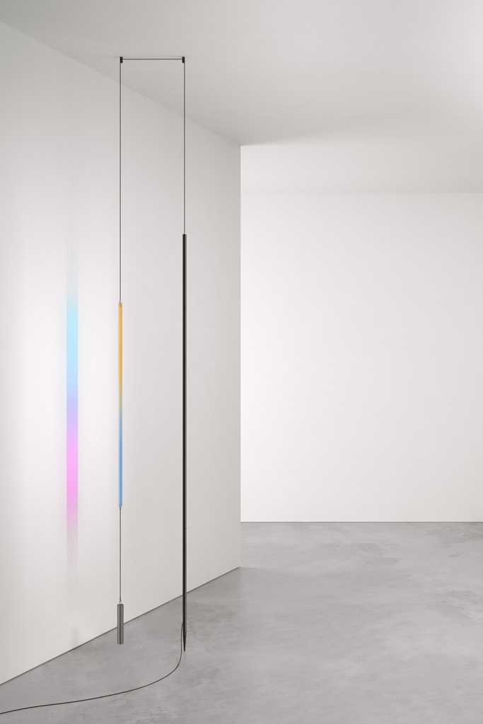 Minimalistic lamp comprised of a slender suspended LED strip facing a suspended rectangle of rainbow-tinted glass to form a long slender three-quarter rectangle. The lamp hangs from the ceiling from a black chord towards the left wall of a stark, all-white room with plain grey linoleum flooring. The rainbow-tinted glass is in the form of a long, slender rectangle on one side of the lamp. Its colors reflect right onto the nearby white wall to its left. The black chord that the glass is attached to is weighed by a small rectangular silver counterweight at the bottom, suspended a few inches from the floor. A long slender black LED strip faces the glass, hanging from the ceiling from the same black chord only a few inches more to the right. A black wire runs from the bottom of the hanging LED light onto the floor and continues out of frame at the bottom left.