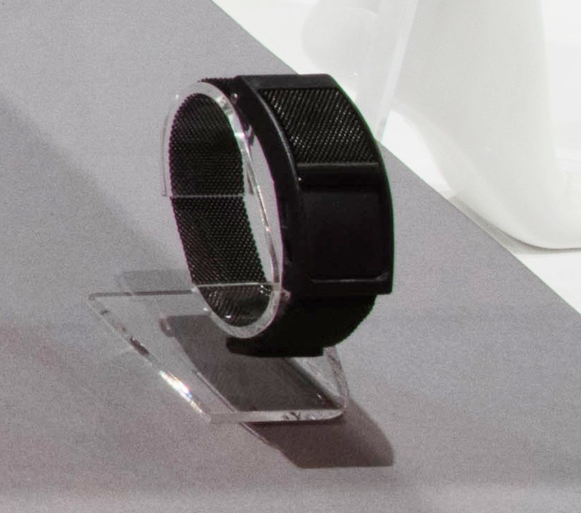 On a clear plastic wrist watch display is an accessibility device with a unique mode if aiding navigation. It is worn on the wrist and provides tactile feedback to its wearer instead of the traditional graphics and audio provided by GPS systems. This is achieved through patterns of vibration. The device takes the shape of a band and is in an all black color. It has a mesh chain for its strap. It is similar to a watch in terms of design construction but is replaced with the technological component of the vibrating device instead of the face of a wristwatch. It is made out of resin, steel, and plastic.