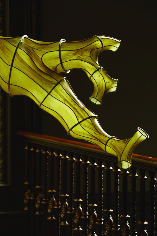 A photograph of a life sized yellowish-green seaweed installation. It is banded and held together by black hoops and lines with two tentacle like extenstions. It extends from the left side to the right side of the photograph, seeming hanging off a higher floor landing, reaching to a darkened room banister.