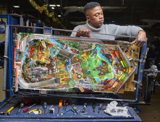 Photograph, Joseph Adeleke testing a pinball machine playfield to ensure all coils, switches, and lamps are working properly