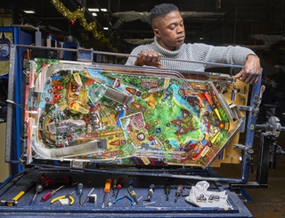 Photograph, Joseph Adeleke testing a pinball machine playfield to ensure all coils, switches, and lamps are working properly