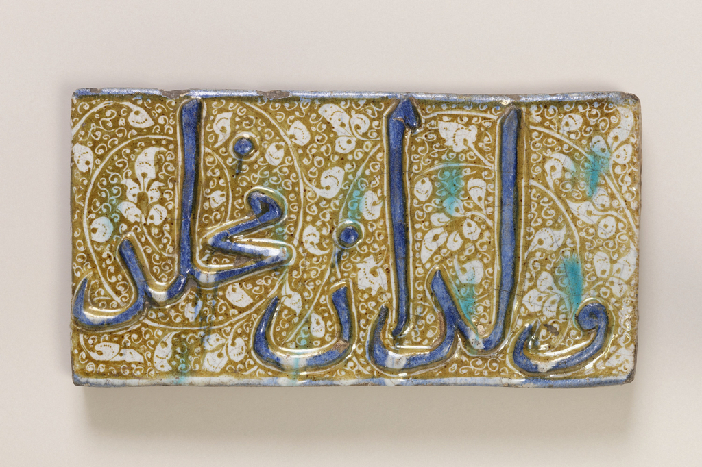 Rectangular tile with raised turqoise inscription on yellow and white scrolled lustered ground. The background is a golden-yellow color with white vines and floral patterning. Sporadic stains of turquoise spots lay on the surface.