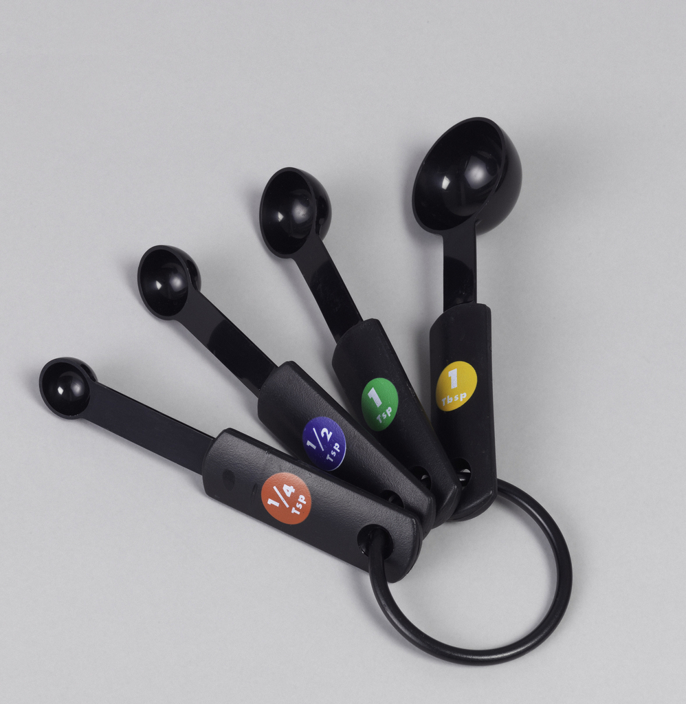 A measuring spoon set with four black spoons of different sizes, each with a circular colored label indicating its measurement, red for a quarter teaspoon, dark blue for a half teaspoon, green for one teaspoon and yellow for one tablespoon. The measurements are printed on each spoon in white inside the corresponding colored circle, and all the spoons are held together on a black plastic ring.