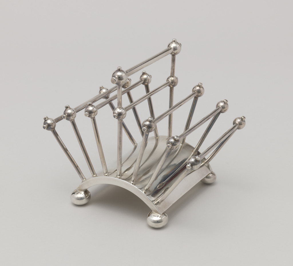 Silver-plated geometric toast rack with an arched base, and upright rectangular handle with three angled rectangular frames on each side. The overall shape is like a book laid on its spine, with the pages fanned out. The base has four bun feet, like flattened balls, one at each corner. The corners of each frame are ornamented with round balls with little nubs protruding, continuing the lines of the vertical and horizontal posts. The handle is taller than the flanking frames, with an additional, thicker horizontal brace at the top, and another, larger pair of ball joints at the top corners.